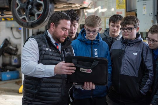A group of young boys in an automotive workshop looking at a diag-box their teacher is holding