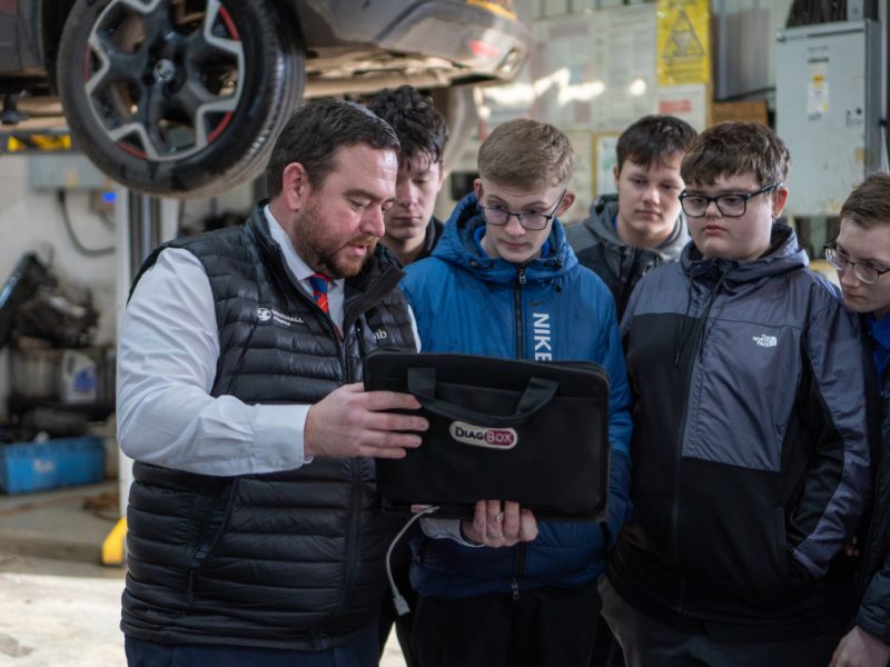 A group of young boys in an automotive workshop looking at a diag-box their teacher is holding
