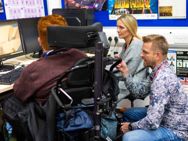 A student sat at a computer in conversation with an employee as part of their work experience placement